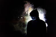 A man in a hooded sweater wears a glowing LED mask against a smoky, night backdrop.