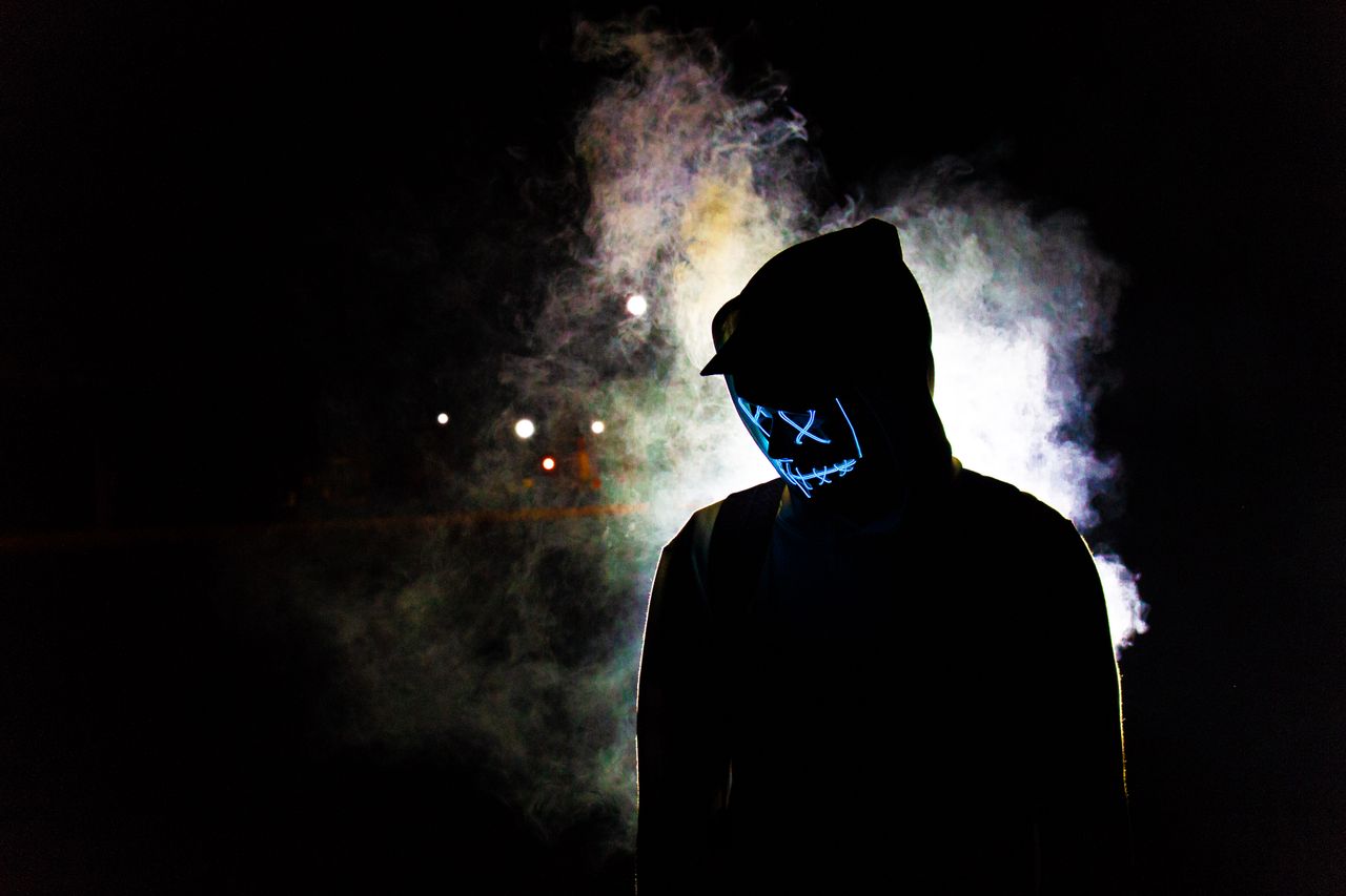 a man in a hooded sweater wears a glowing led mask against a smoky nig