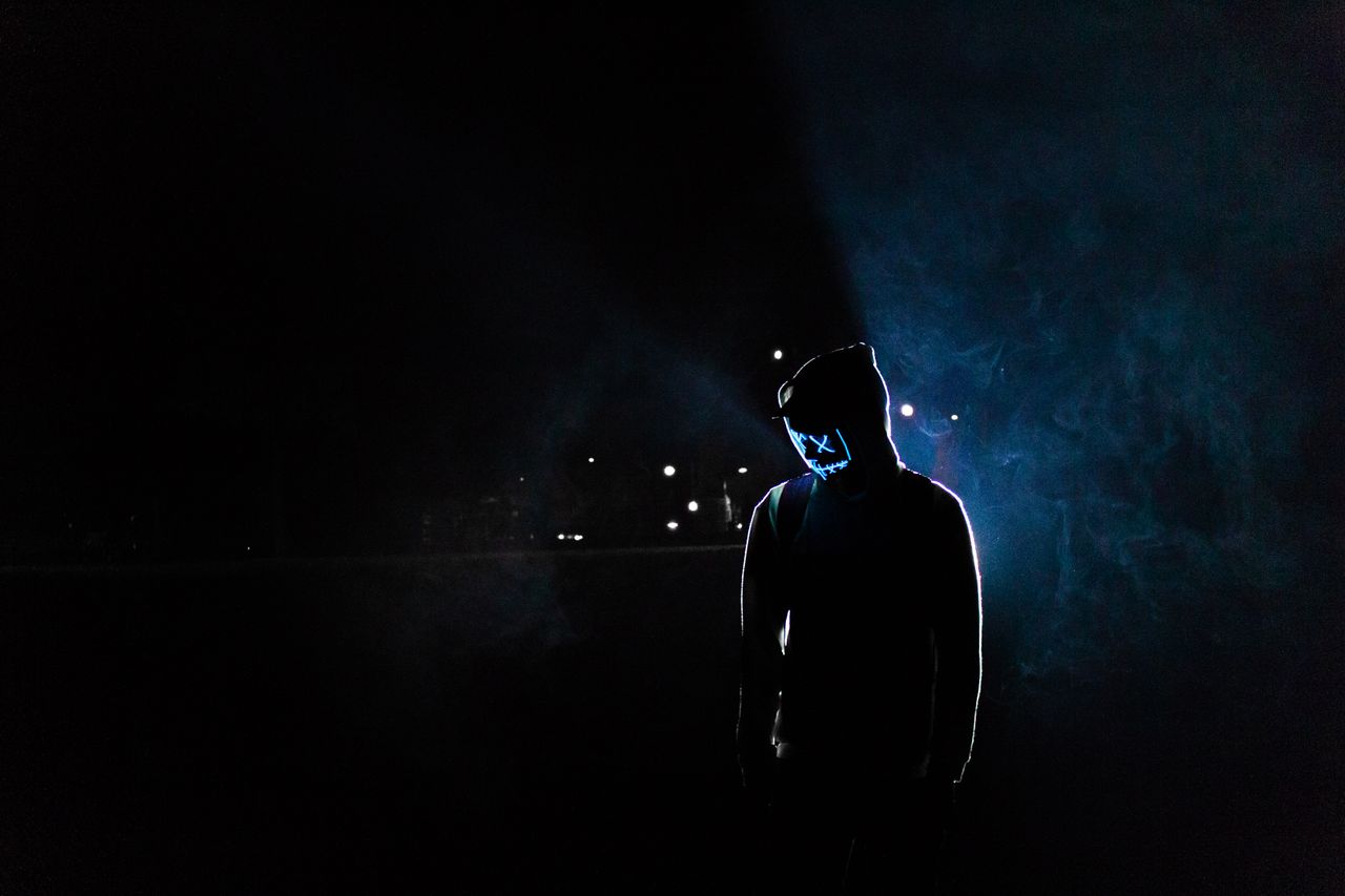 silhouette of a man in a glowing led mask and hoodie in a dark foggy s