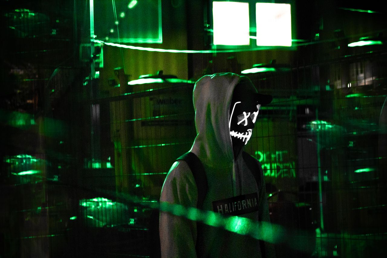 man in a hoodie with glowing mask amidst neon green lights evoking mys