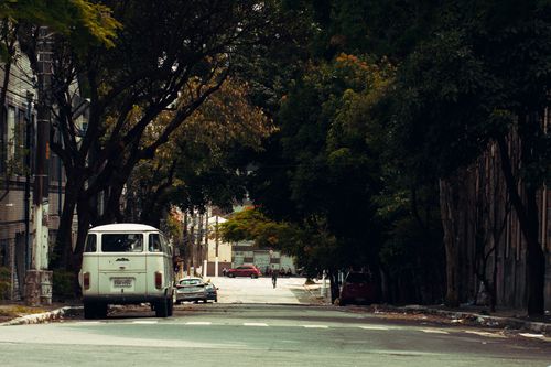 Free A tranquil city street lined with trees, featuring parked cars and a classic van. Stock Photo