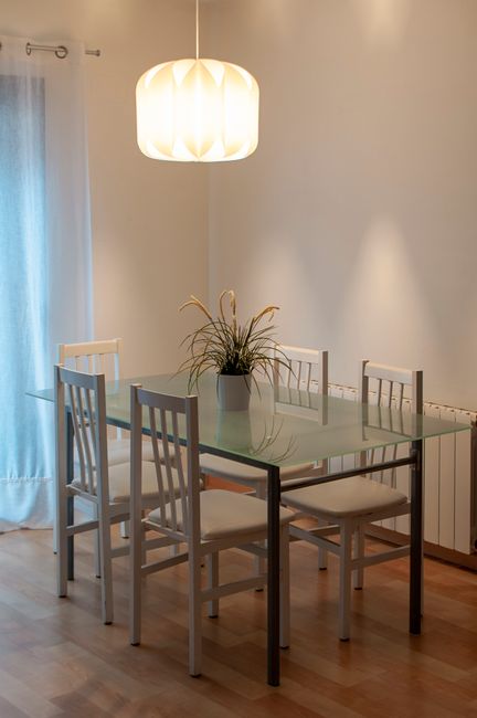 Fotomural Comedor elegante con mesa de cristal, planta en maceta e iluminación moderna.