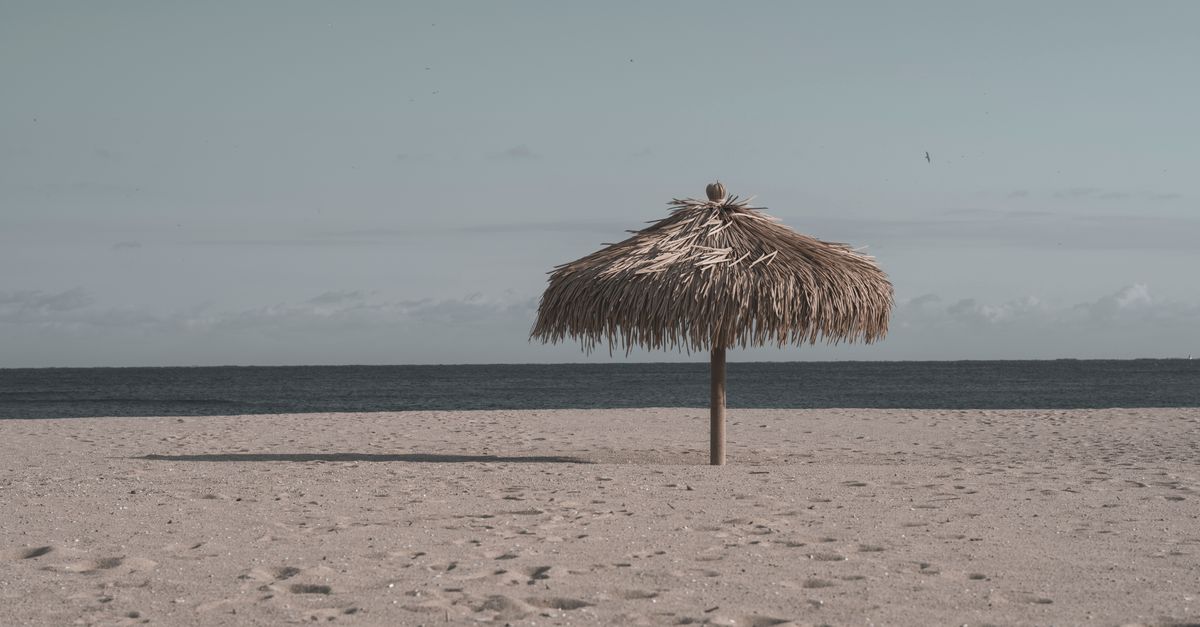 Peaceful sandy beach with a single straw umbrella under a blue sky and open ocean.