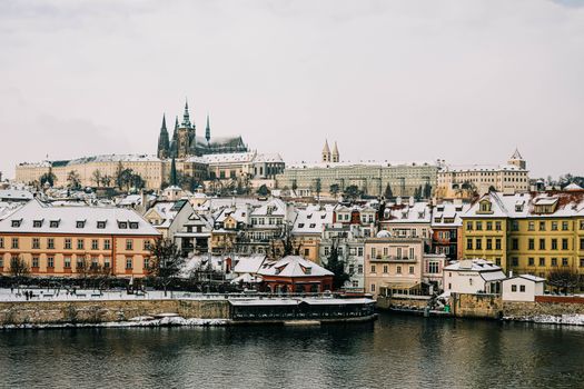 Explore a stunning winter view of Prague with snow-covered rooftops and historic architecture along the river.