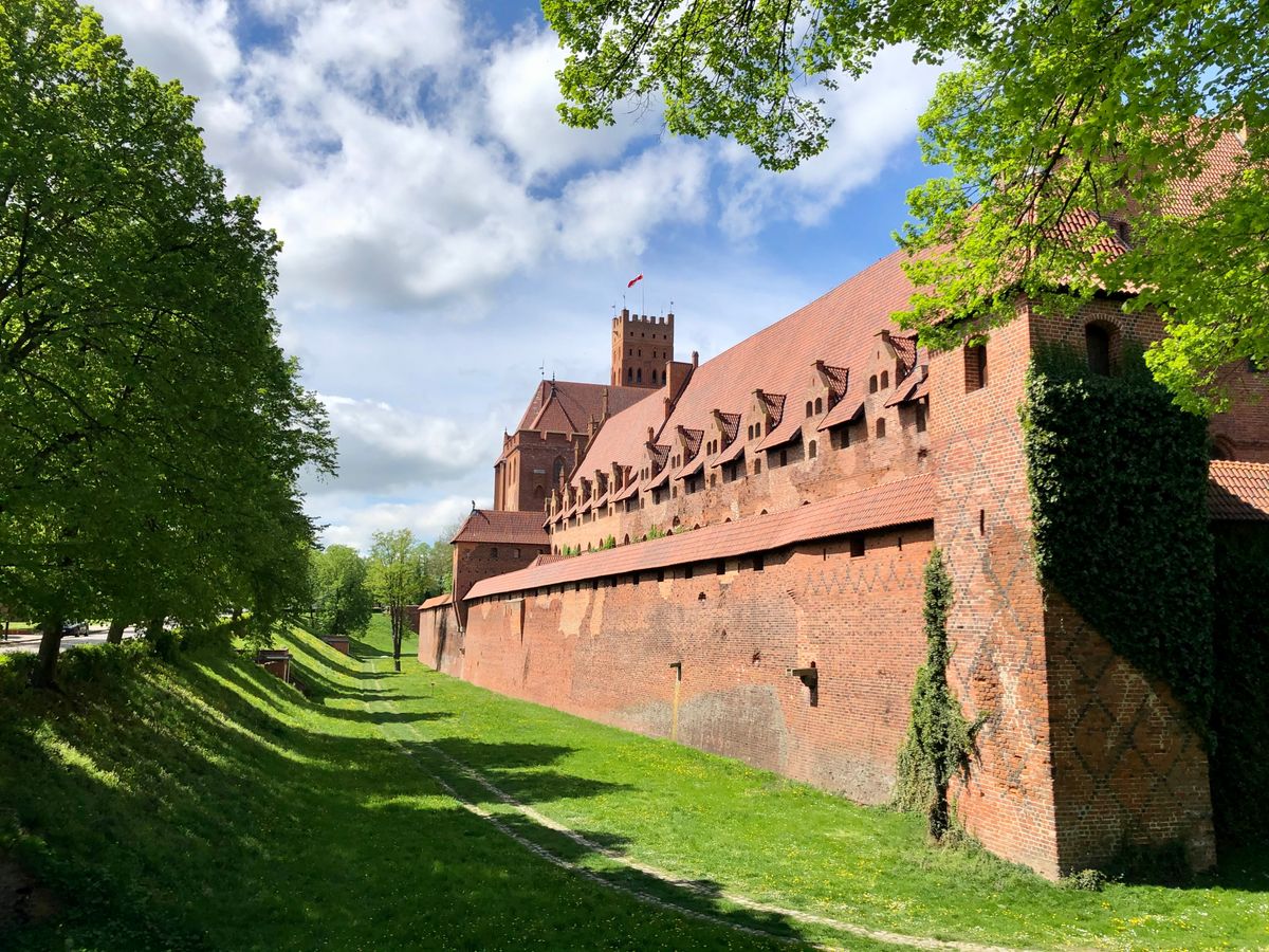 A long red-brick outer wall running along a grassy ditch in strong sun, ivy climbing its corner over a diamond lattice of darker bricks, with the castle's roofs and a tall tower beyond.