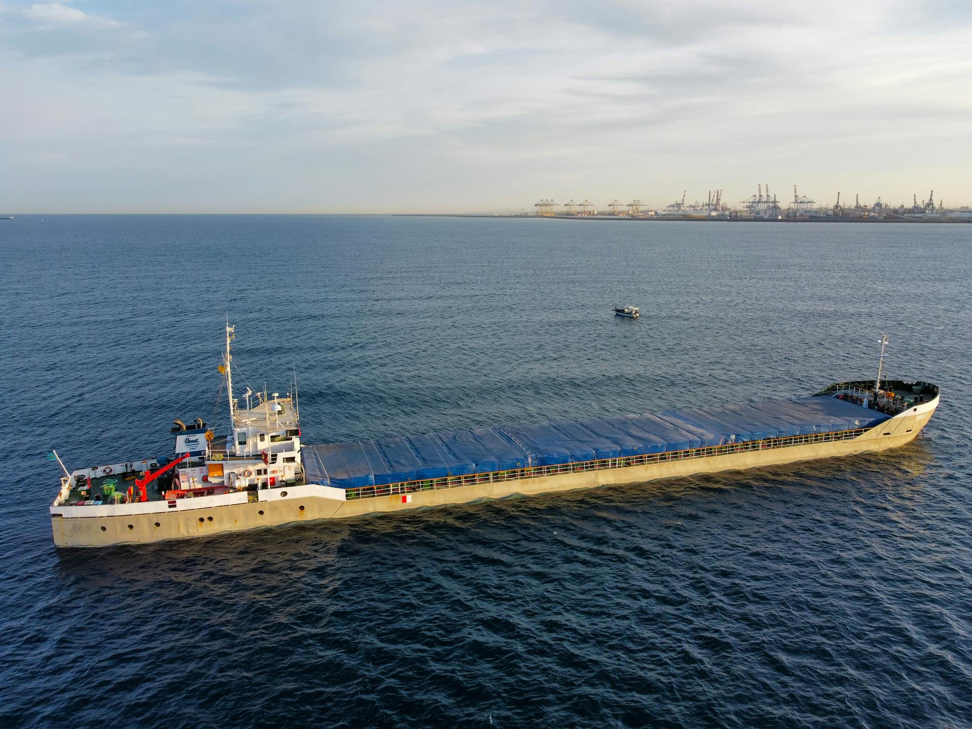 Aerial view of a cargo ship sailing near a bustling industrial harbor with cranes in the background.
