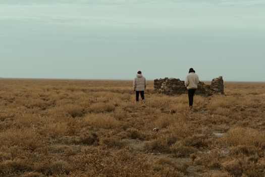 Two people wander a vast open field with ancient stone ruins under a cold sky.
