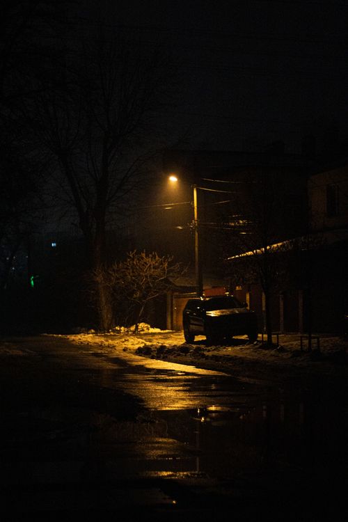 Free A moody, dimly lit street in Ukraine featuring a parked car under streetlights and snowy surroundings at night. Stock Photo