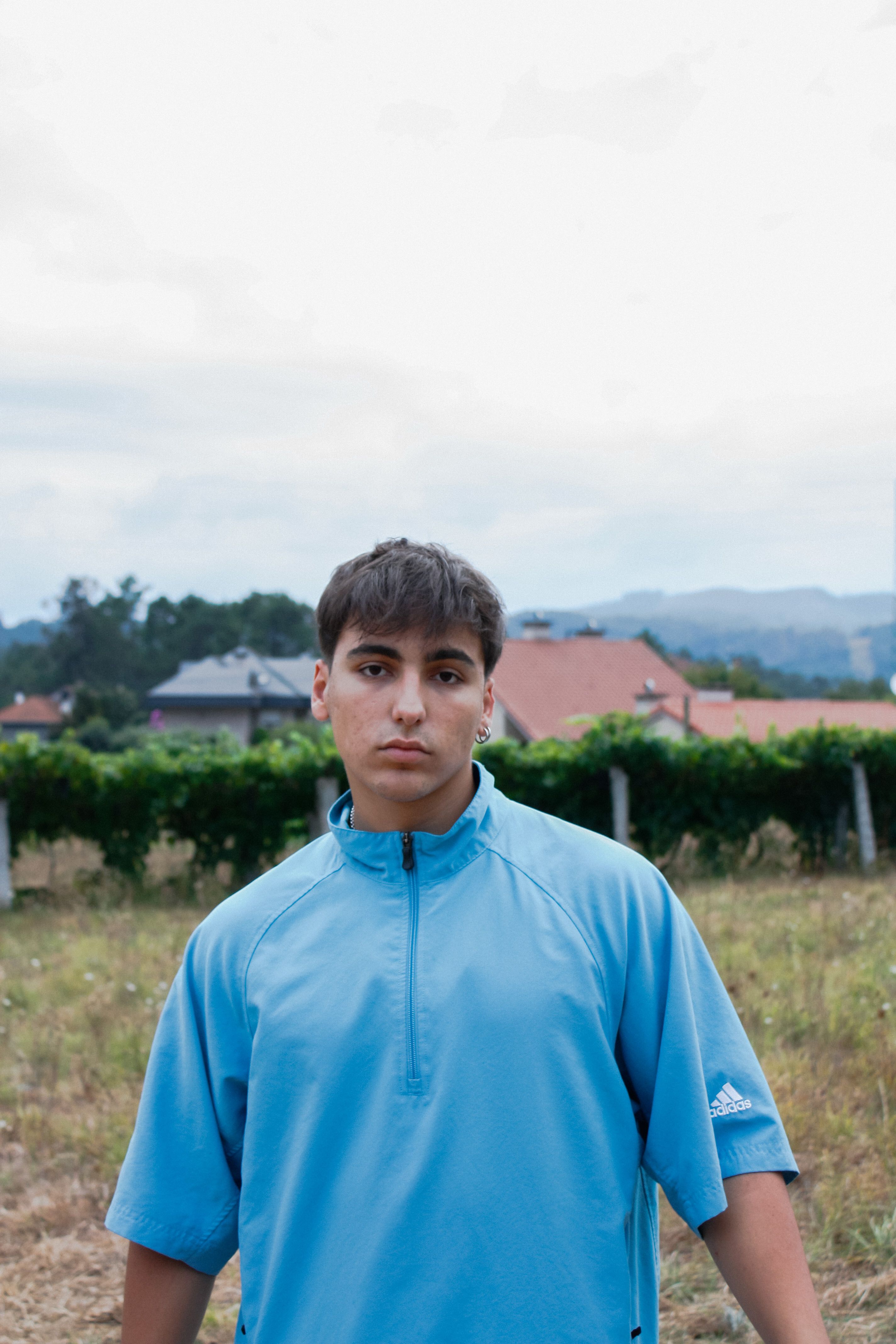 Portrait of a young man with short hair standing in a rural outdoor setting wearing a blue shirt.