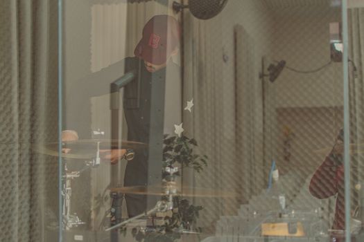 A man in a soundproof studio playing drums, captured creatively behind a glass panel.