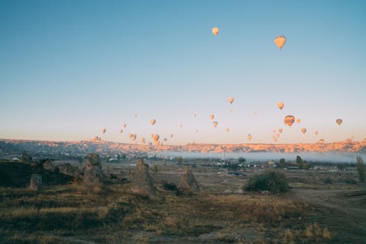 Scenic vista of numerous hot air balloons soaring above a rugged landscape during sunrise.