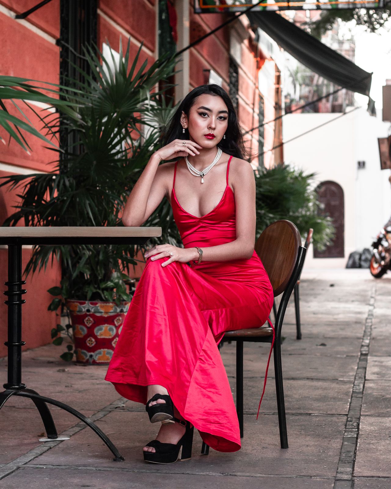 Woman in a red satin slip dress with black platform sandals sitting at an outdoor cafe table