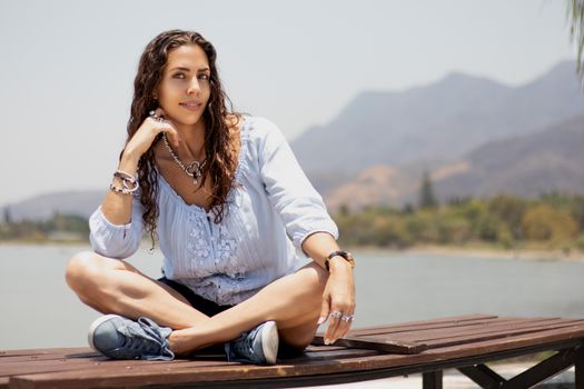 A woman with curly hair sitting on a wooden bench by a lake, enjoying a sunny day.