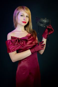 Stylish woman in a red satin gown poses with a black carnival mask against a dark background.