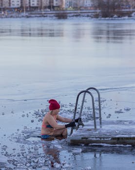 A woman wearing a swimsuit and hat takes a dip in a frozen lake by a ladder. Cold winter activity.