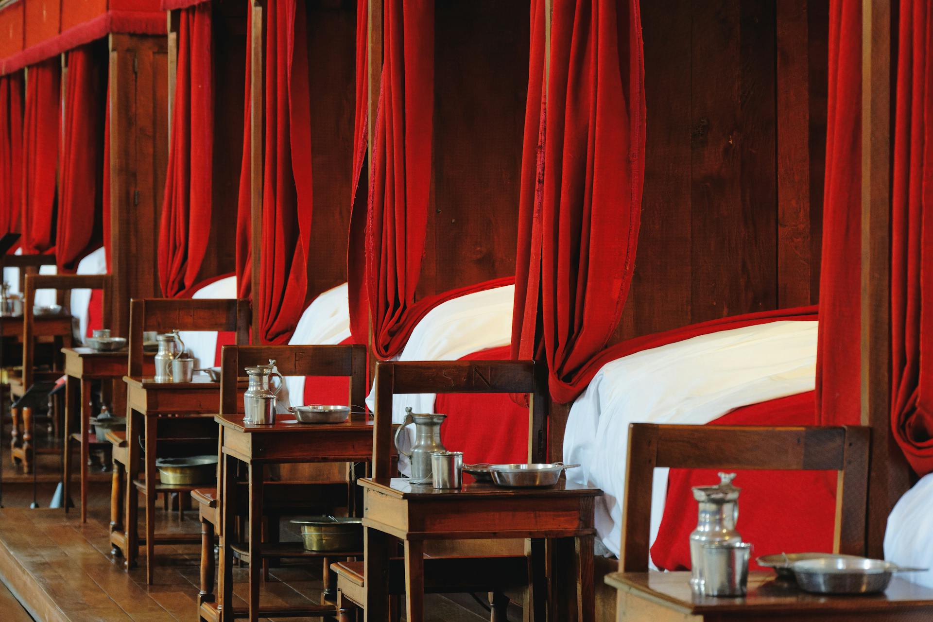 Interior of a historic hospital ward in Vougeot, showcasing red-curtained beds and antique furniture.