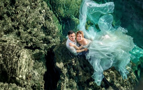Free A romantic couple embraces by a clear rocky shore in flowing attire under sunlight. Stock Photo
