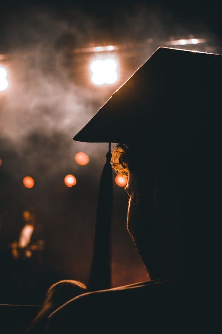Silhouette of a graduate with a mortarboard under bright stage lights, creating a dramatic ambiance. Silhouette of a graduate with a mortarboard under bright stage lights, creating a dramatic ambiance.