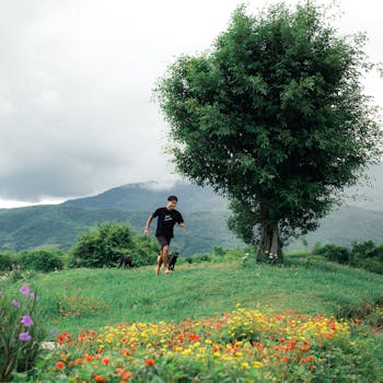 A man runs with a dog in a vibrant meadow with flowers and a tree, set against mountain scenery.