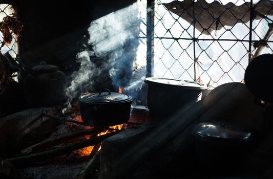 A traditional cooking setup with pots over a flame, emitting smoke in a dark rustic kitchen.