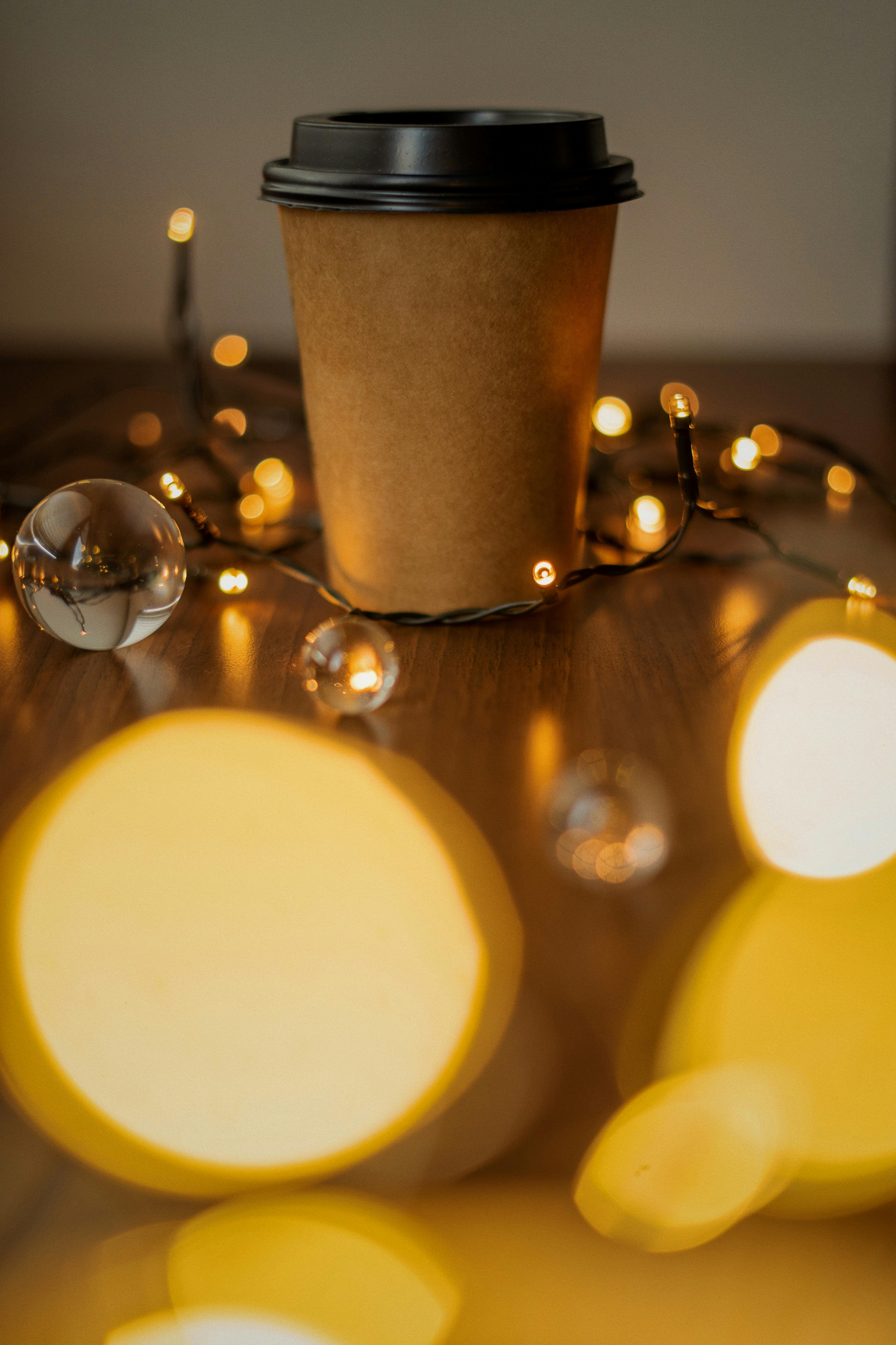 Paper coffee cup surrounded by warm, glowing bokeh lights on a wooden table, creating a festive atmosphere.