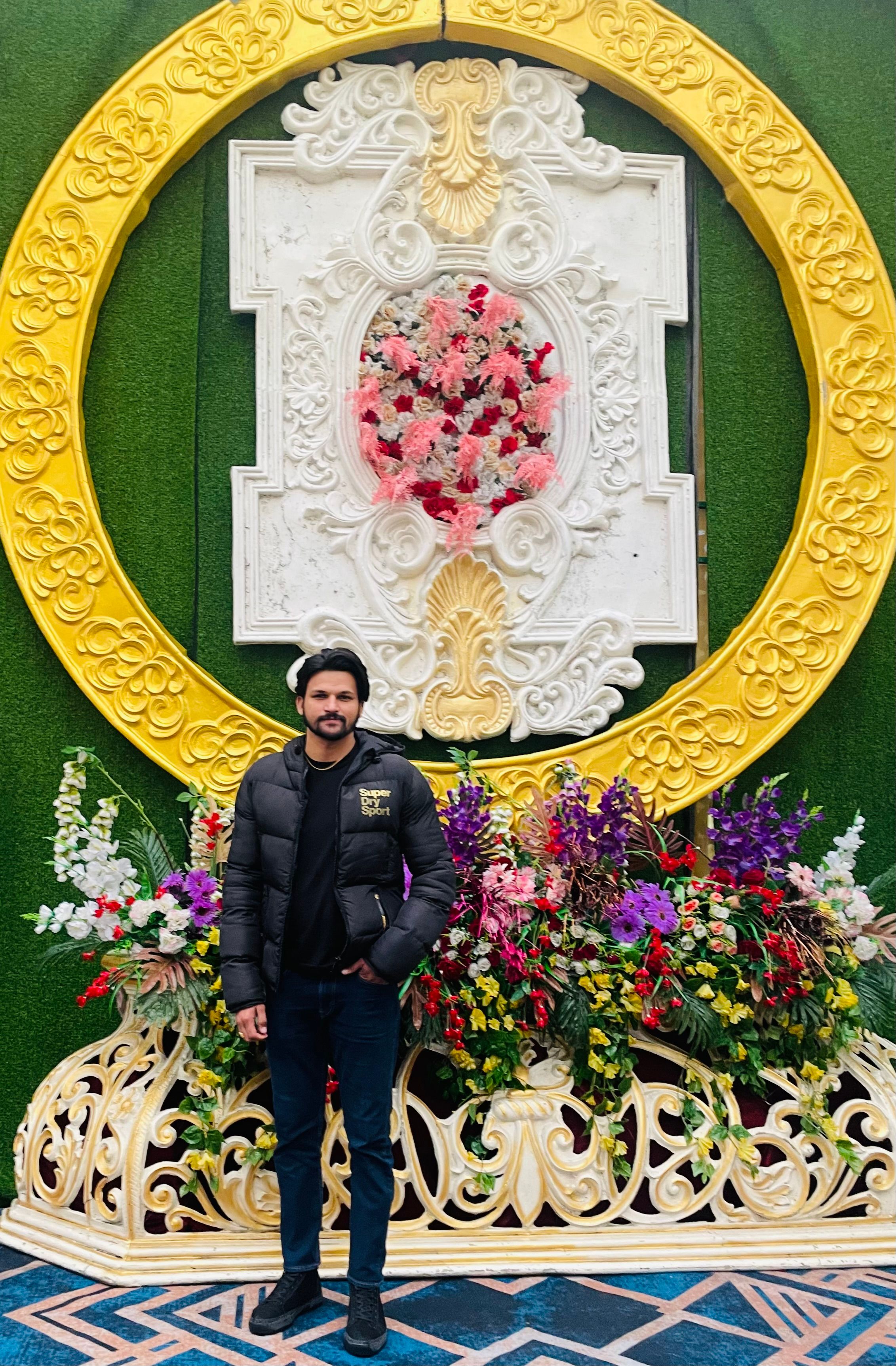 A man poses in front of an ornate floral wall decoration with lush colors.