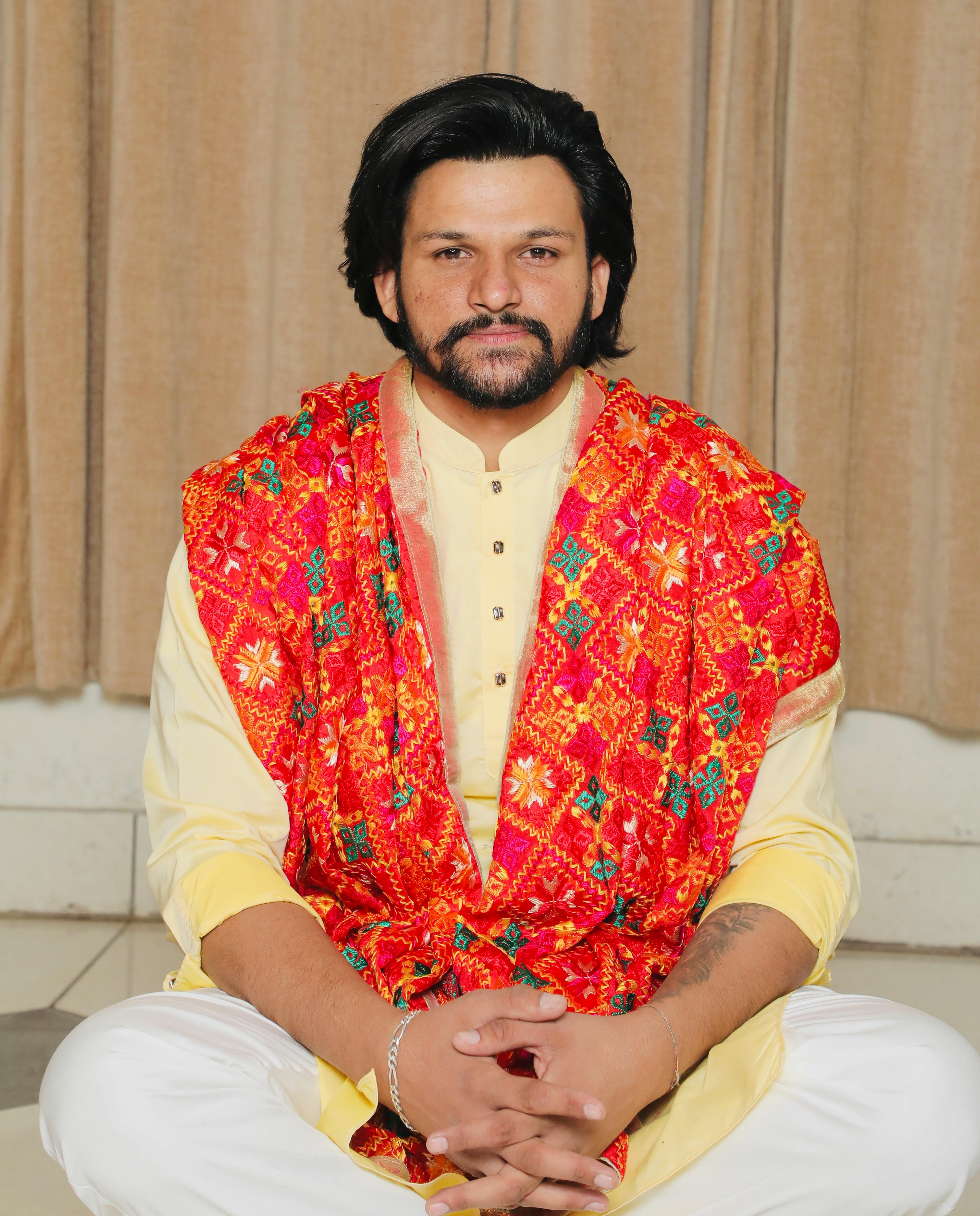 A man with facial hair wearing traditional colorful attire, sitting indoors.