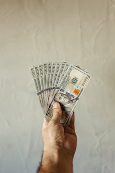A hand displays a fan of US hundred dollar bills against a plain background.