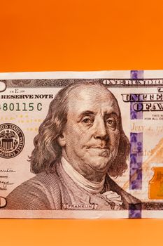 Close-up of a one hundred dollar bill featuring Benjamin Franklin against an orange background.