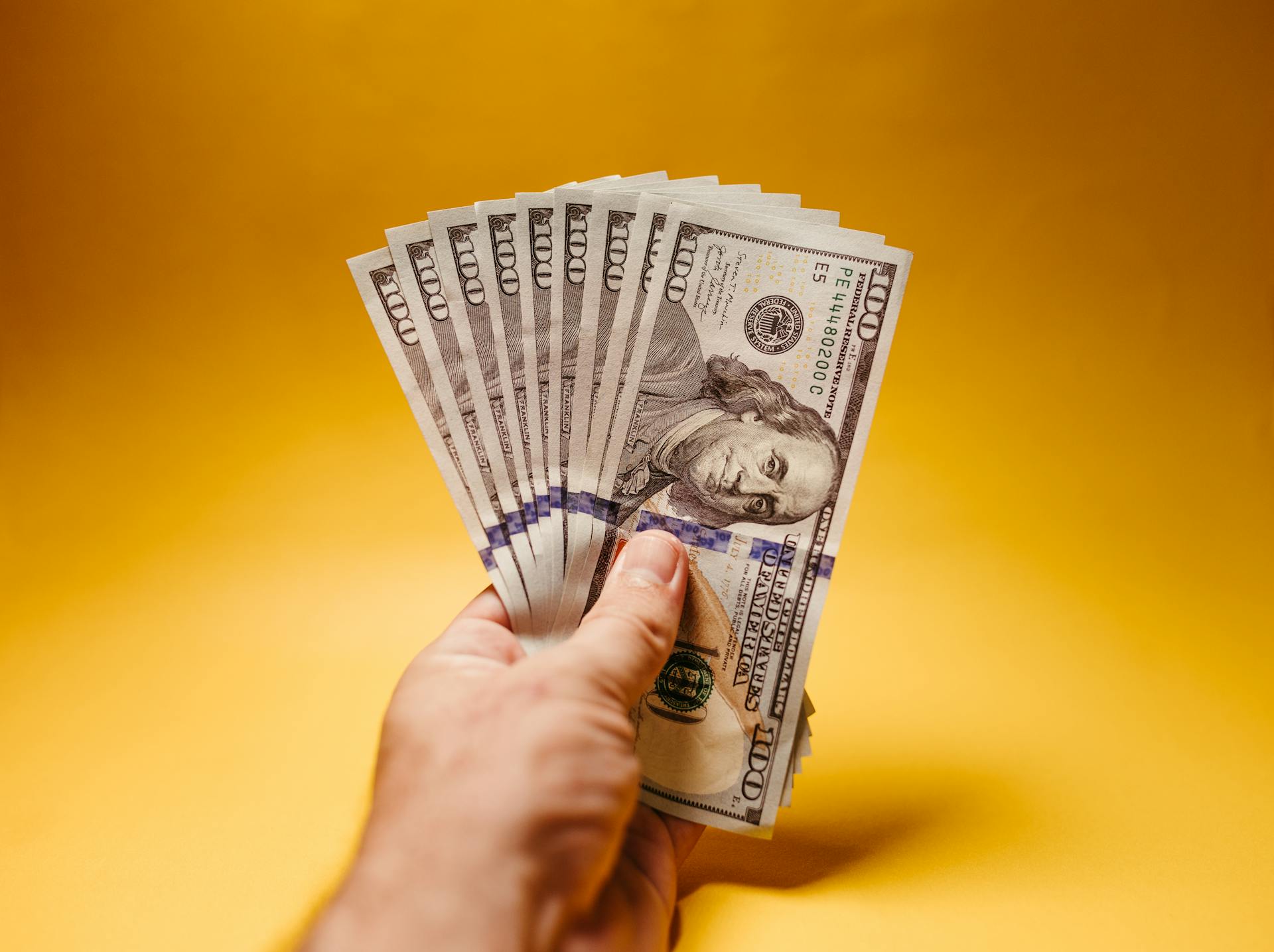 Close-up of a hand holding US hundred dollar bills against a vibrant yellow background.