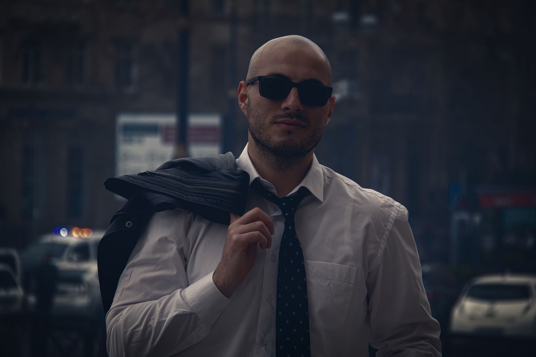 A confident bald man in sunglasses and necktie stands outdoors, exuding urban style.