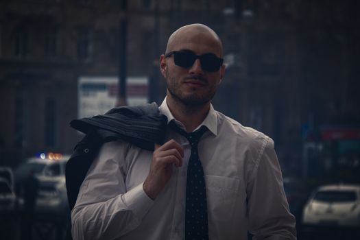 A confident bald man in sunglasses and necktie stands outdoors, exuding urban style.