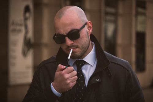 Free Bald man with sunglasses lights a cigarette outdoors, portraying a confident demeanor. Stock Photo