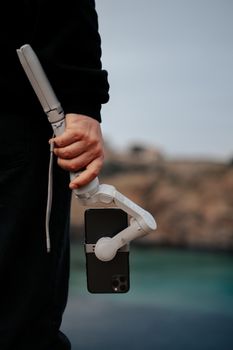 Close-up of a person holding a smartphone stabilized by a gimbal, set against an outdoor background.