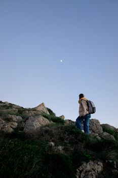 A solo hiker with a backpack ascends rocky terrain under a clear moonlit sky.
