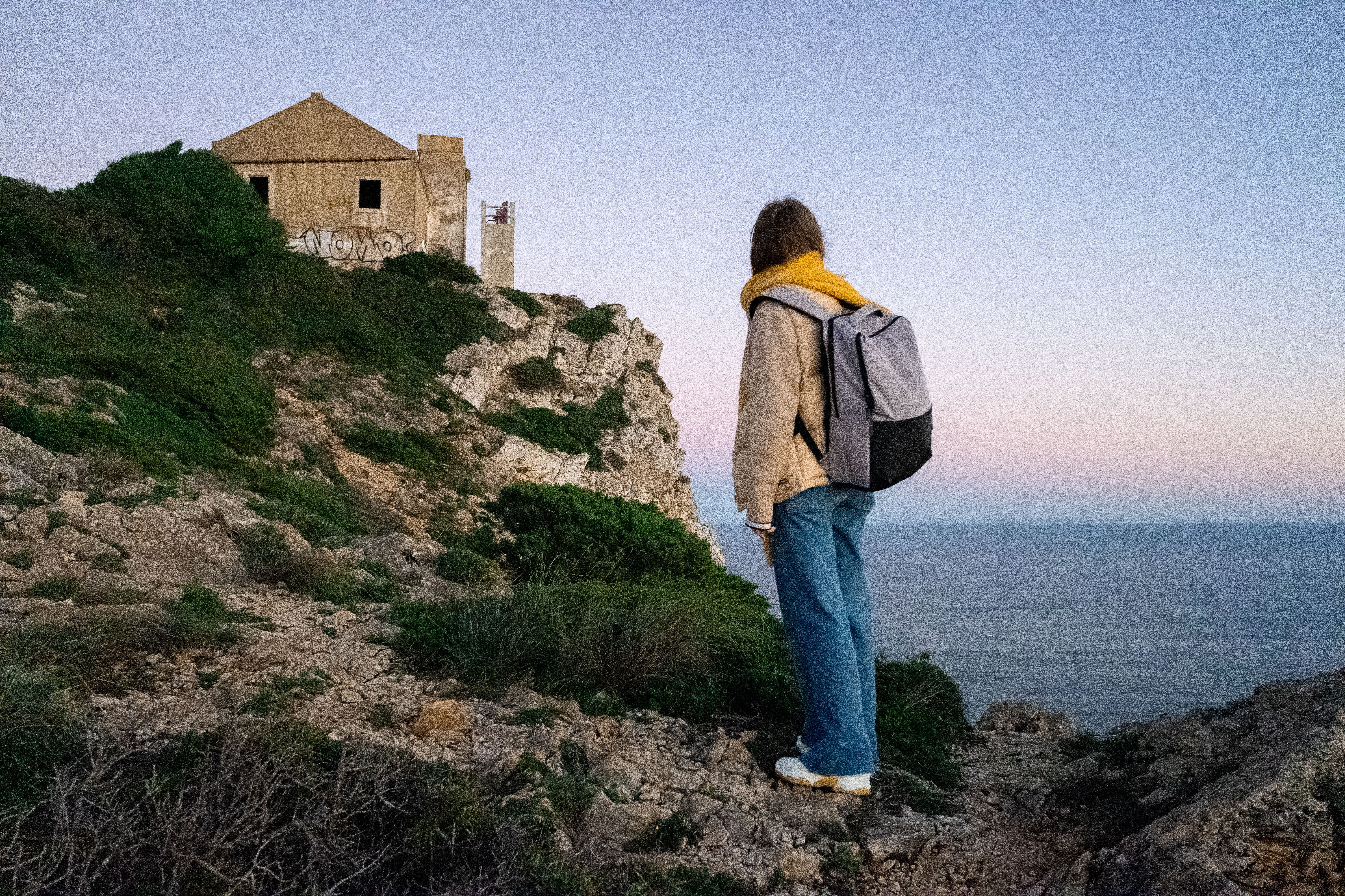 A backpacker gazes at an abandoned house perched on coastal cliffs during sunset.
