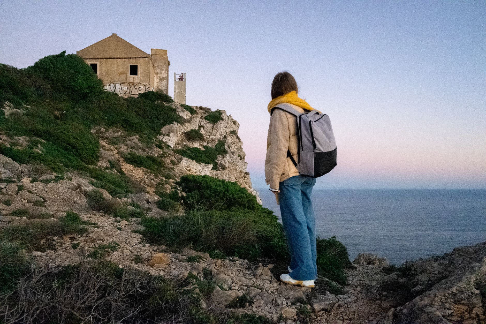 A backpacker gazes at an abandoned house perched on coastal cliffs during sunset.