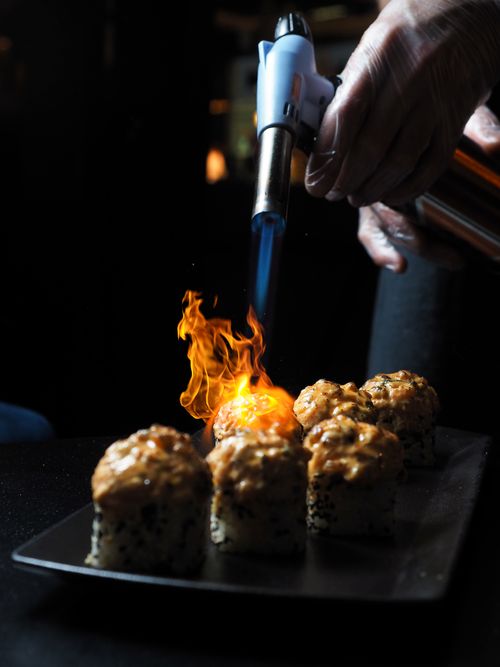 Free A close-up of a chef using a blowtorch to prepare gourmet sushi rolls in a dimly lit restaurant setting. Stock Photo