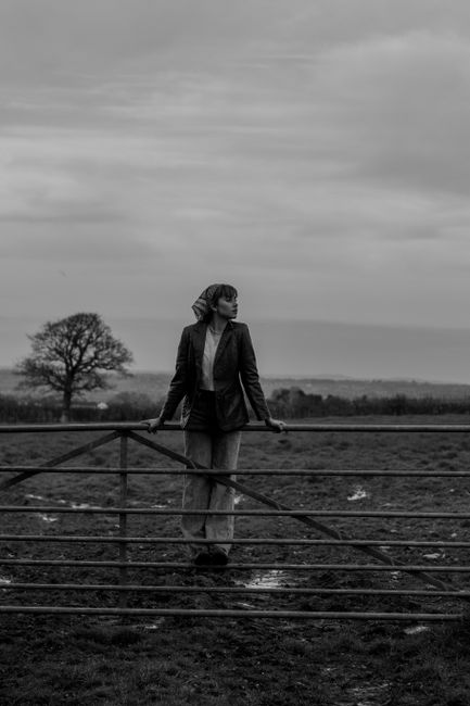 A woman leans on a fence in a rural landscape, captured in black and white photography.