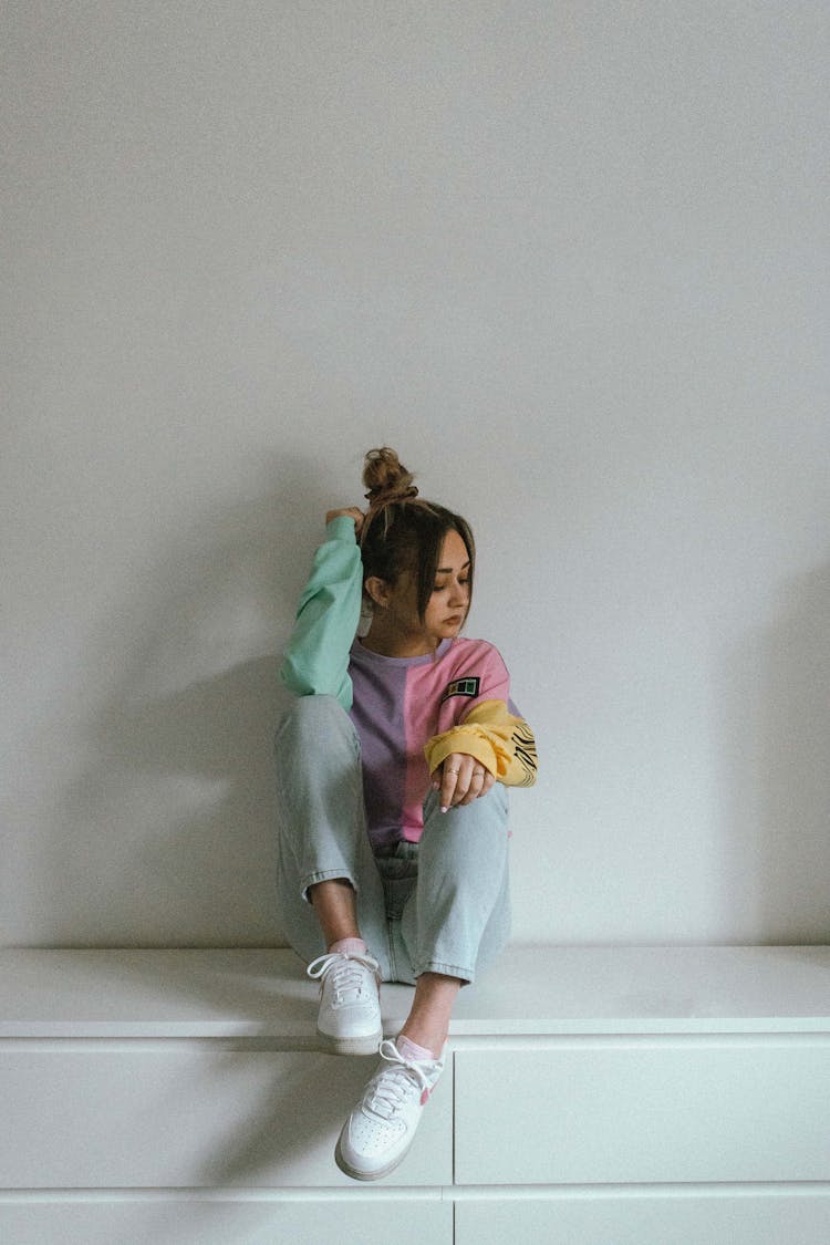 Teenage Girl In Colorful Long Sleeves Shirt And Denim Pants Sitting On A Wooden Cabinet