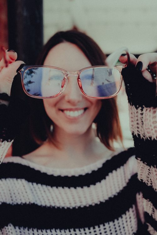 Free A cheerful woman in a striped sweater holding glasses with a blurred reflection outdoors. Stock Photo
