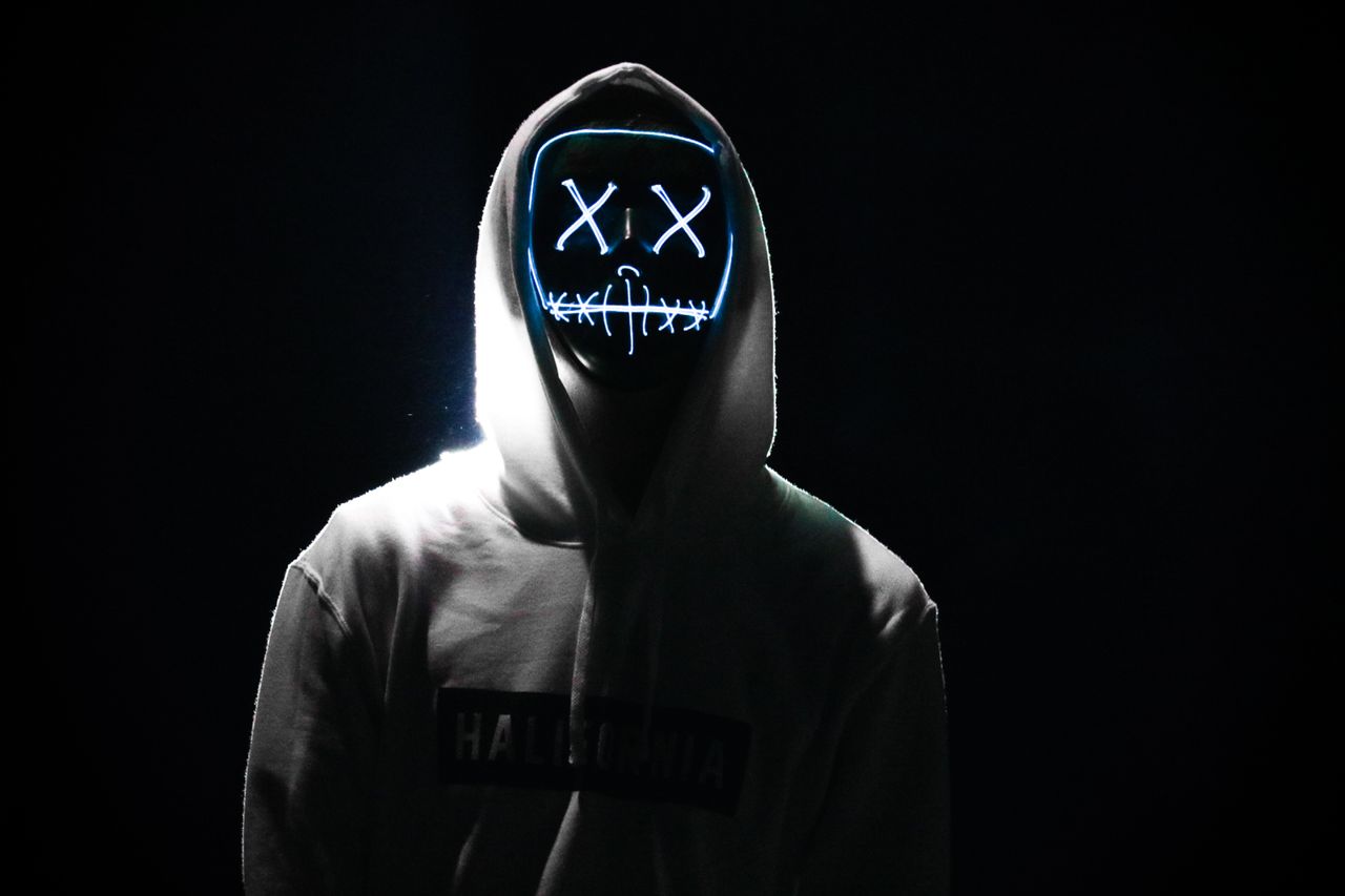a person in a hooded sweatshirt wears a glowing led mask in a dark mys