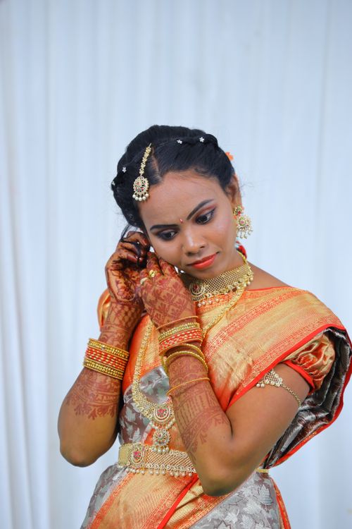 Free A woman in a vibrant orange saree with henna-adorned hands adjusting her earrings. Stock Photo