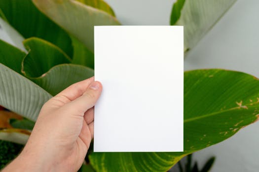 A person's hand holding a blank white paper in front of lush green leaves.