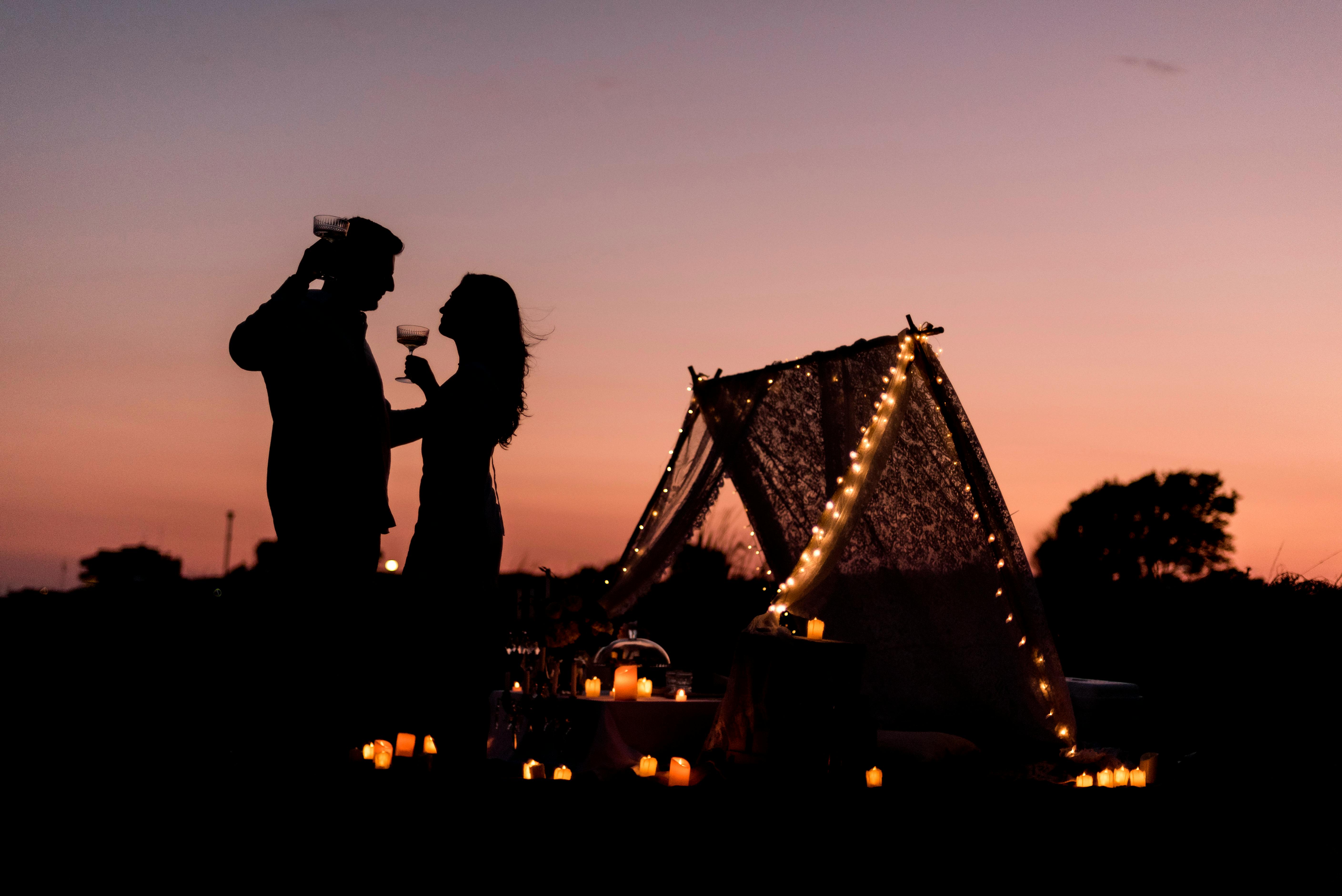 Silhouette of a couple toasting wine glasses during a romantic sunset camping setup with candles.
