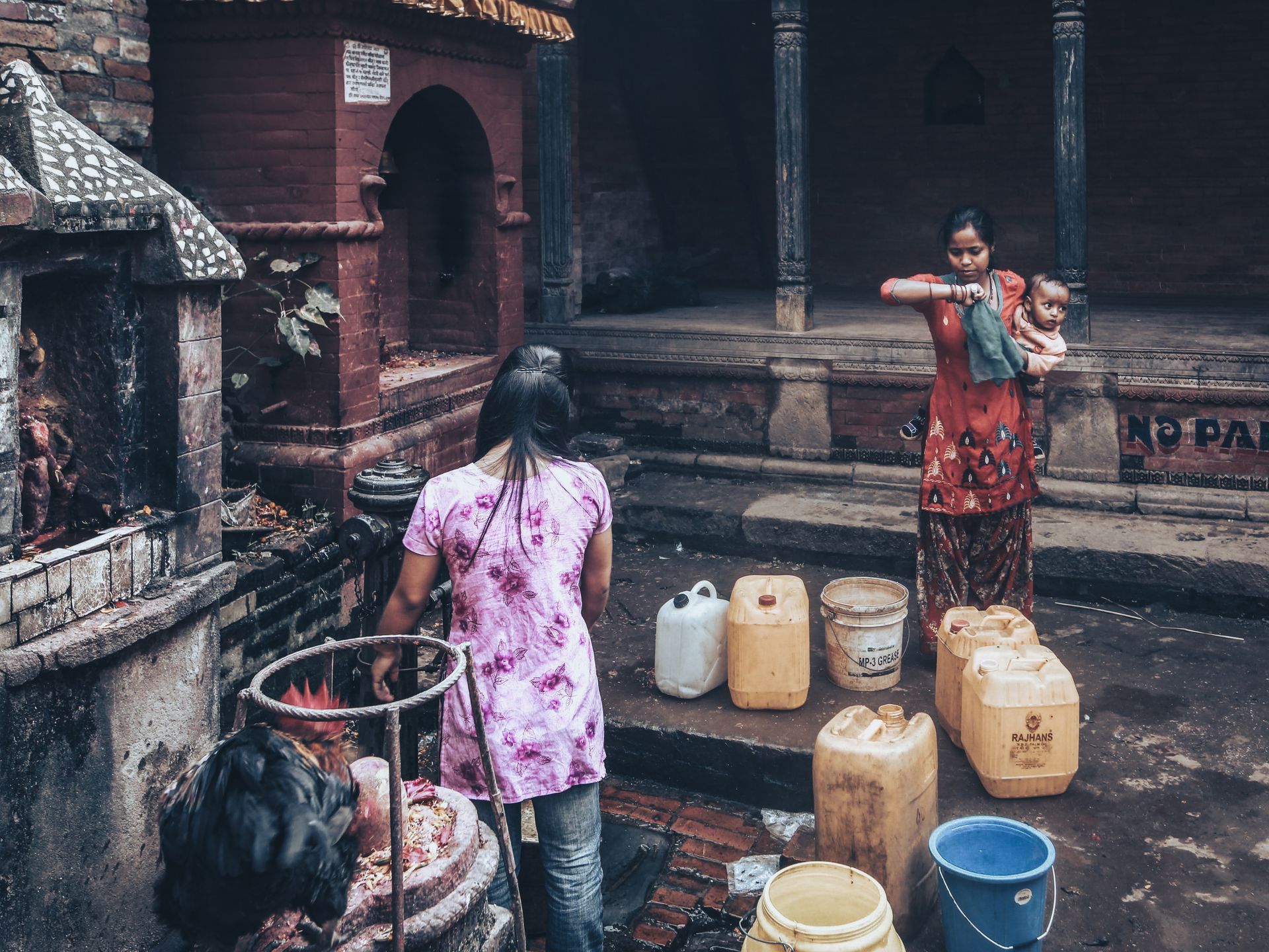 Women fill containers at a water pump station, engaging in daily chores outdoors.