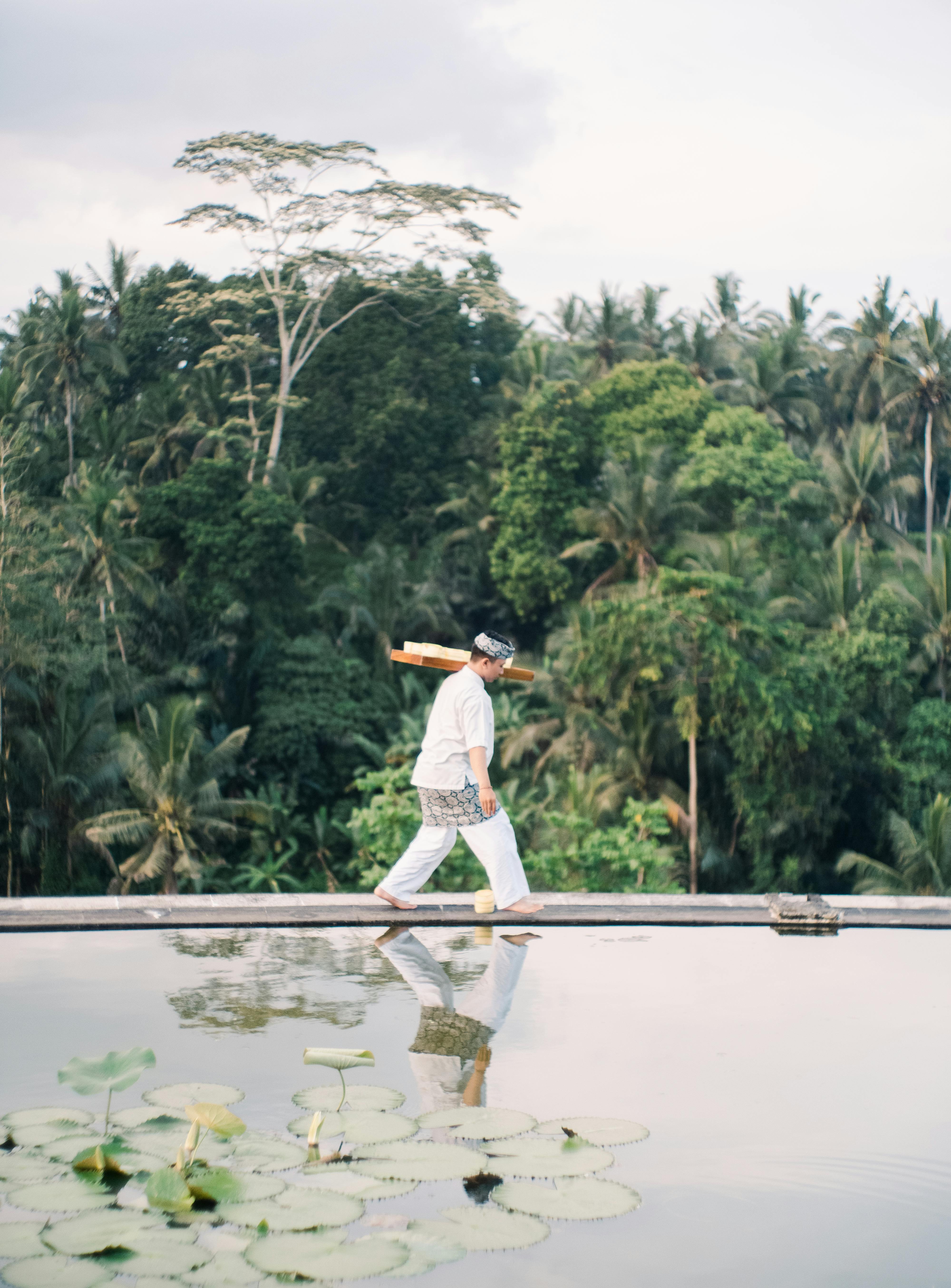 A man in traditional clothing walks by a pond reflecting lush tropical trees.