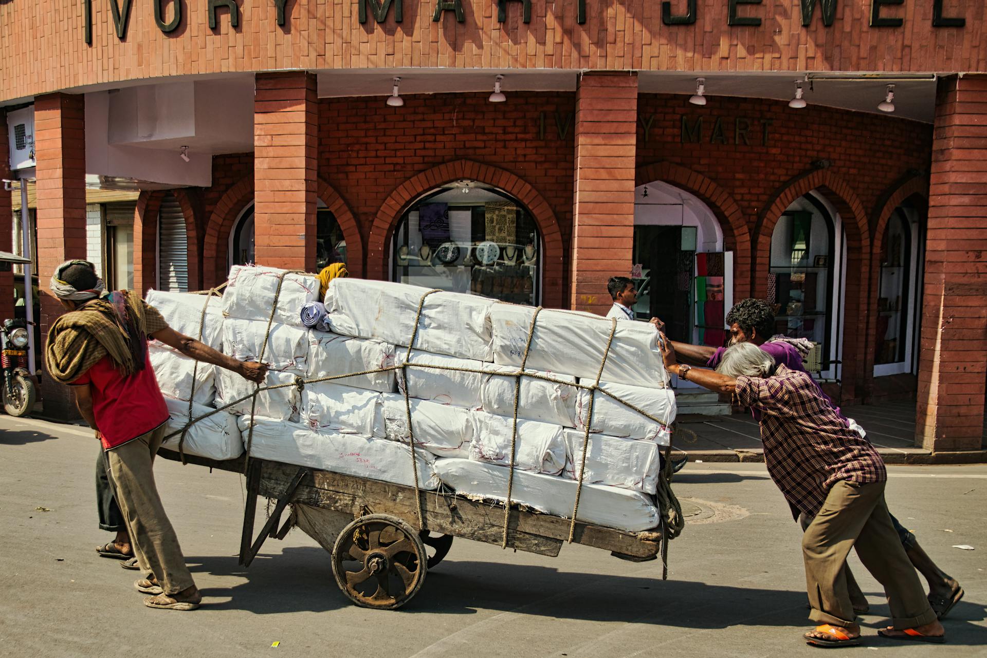 Two men exert effort pushing a heavy cart full of packages on an urban city street.