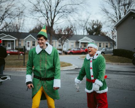 Two adults dressed as elves laughing in a winter neighborhood holiday scene.