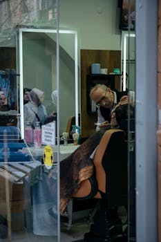 A barber trims a customer's beard inside a cozy barbershop, seen through a glass window.